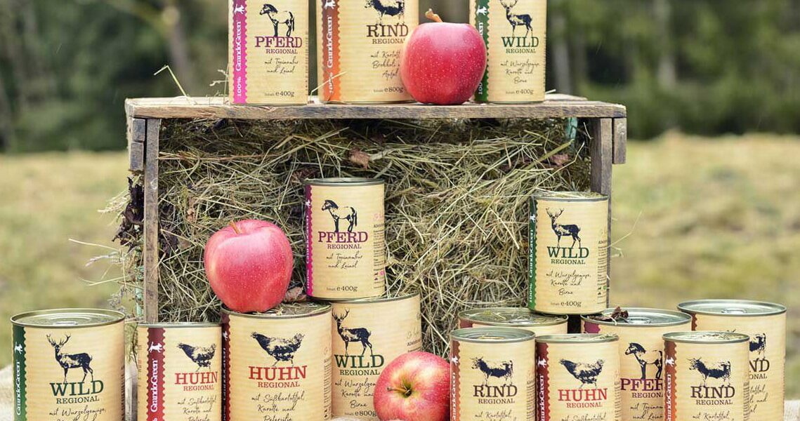 Regionales Hundefutter – Sortenvielfalt Pferd, Rind, Wild & Huhn Mehrere Konservendosen mit regionalem Hundefutter-Sortiment (Pferd, Rind, Wild, Huhn) auf einer Heugabel und am Boden, umgeben von Äpfeln vor Waldkulisse.