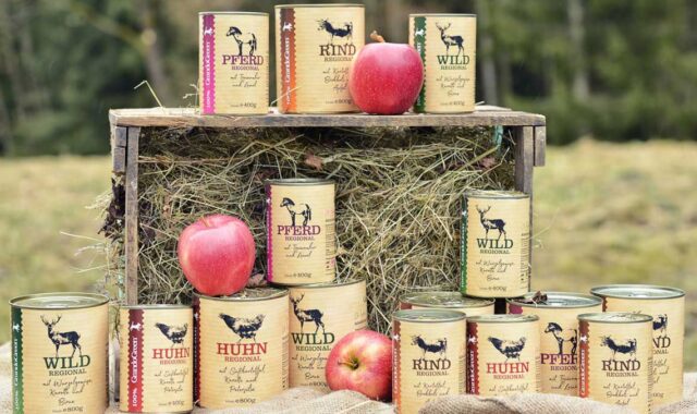 Mehrere Konservendosen mit regionalem Hundefutter-Sortiment (Pferd, Rind, Wild, Huhn) auf einer Heugabel und am Boden, umgeben von Äpfeln vor Waldkulisse.