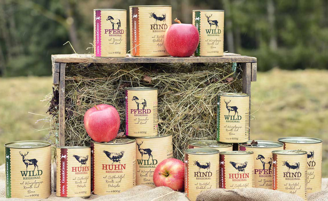 Mehrere Konservendosen mit regionalem Hundefutter-Sortiment (Pferd, Rind, Wild, Huhn) auf einer Heugabel und am Boden, umgeben von Äpfeln vor Waldkulisse.